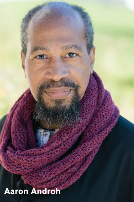 A headshot of actor Aaron Androh, a middle-aged Black man with a receding hairline and with greying hair, beard and mustache. He is wearing a dark shirt and has a highly textured and voluminous raspberry-colored scarf around his neck.