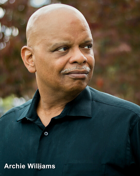 a headshot of actor Archie Williams, a Black man with a shaved head and white pencil-mustache. He is gazing to the right and the photo was shot outdoors against a blurry natural background. He is wearing a dark green collared shirt, open at the neck.