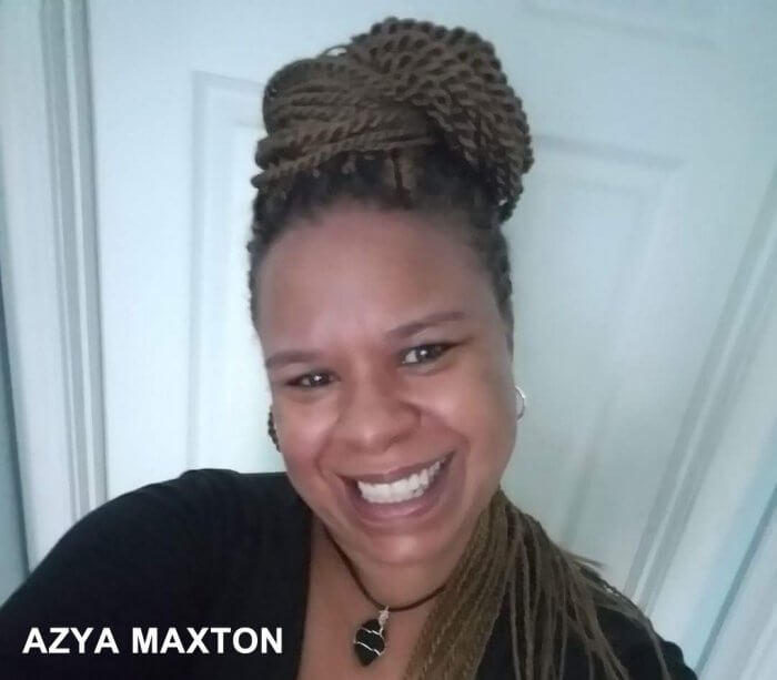 A headshot of actor Azya Maxton, a middle-aged Black woman looking at the camera with a big smile on her face. She has long braids which are coiled on top of head and winding down onto her left shoulder. She is wearing a black shirt and a necklace with a black stone wrapped in gold wire hanging from a leather thing.