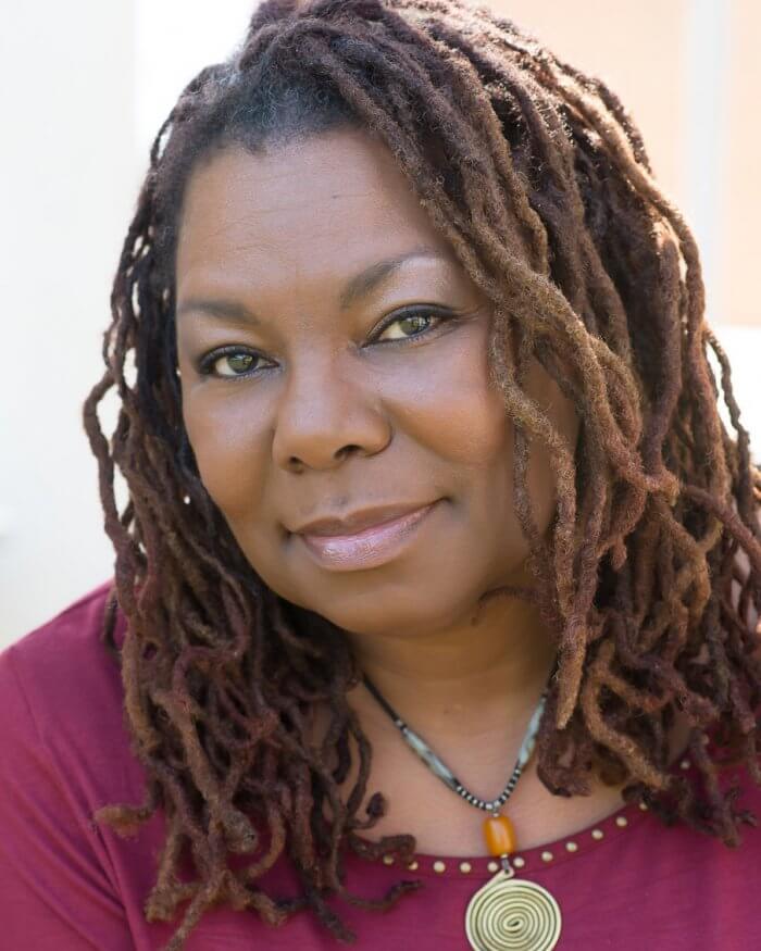 Photograph of Deletta Gillespie, a middle-aged Black woman looking at the camera with a slight smile. She has shoulder-length dreads, and is wearing a necklace of grey and amber beads with above a bronze spiral. She is wearing a raspberry-colored shirt with gold bead edging around the scoop neck.