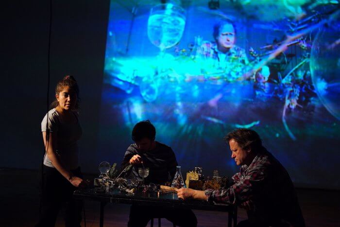 A rehearsal of Serious Play's "Moving Water" showing 3 actors in front of a projected image in blues, aquas and purples. A wavy, watery image, it shows one of the actors faces at the top of the frame. In front of the projection an actress stands left of a table looking out at us. She is lit from the left side. The rest of her body is shadowed. She is next to a table filled with glass paraphernalia. A man is is seated behind the table in shadow pouring water into a glass. A 3rd man sits at the table opposite the woman. He is lit from the left and we can see that he is reading a piece of paper.